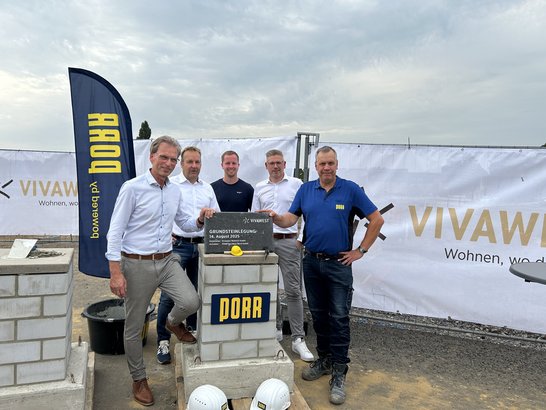 Das Bild zeigt eine Gruppe von fünf Personen bei einer feierlichen Grundsteinlegung. Sie stehen hinter einem Betonblock. Eine Person hält eine Plakette. Vor dem Betonblock liegen zwei weiße Bauhelme.  Im Hintergrund sind ein Banner sowie eine Fahne zu erkennen. Die Szenerie spielt sich auf einer Baustelle im Freien unter einem bewölkten Himmel ab.
