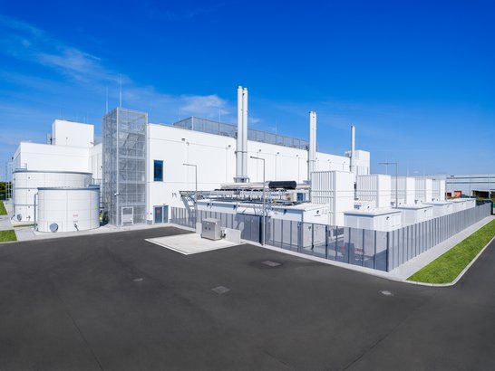 The image shows a modern industrial building with several large white structures and chimneys. In the foreground is a large paved area. Technical equipment such as tanks and pipes are visible at the side of the building. The architecture appears clean and tidy, and the background shows a blue sky, giving the image a bright and uncluttered atmosphere.
