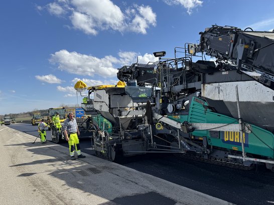 Bild zeigt Geräte des Verkehrswegebaus, daneben ein Mitarbeiter in Schutzkleidung