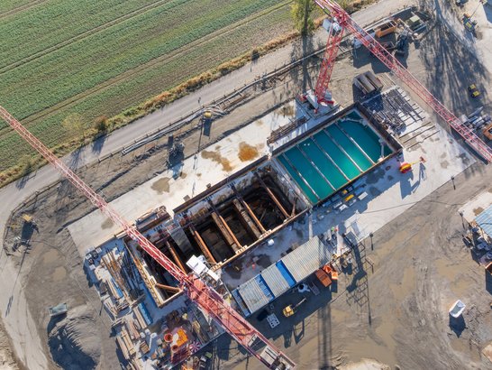 The image shows the excavation pit from above. You can see that the right-hand side is filled with blue water. Construction machinery and cranes are positioned around it.