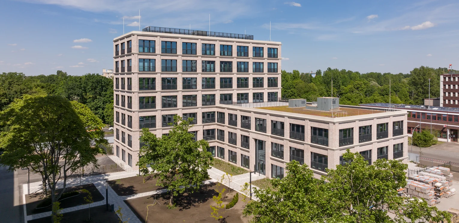 TechnoCampus, Berlin. Picture of the new building with extension; the façade has many large rectangular windows; in the foreground there are trees on the forecourt; in the background you can see a forest; the sky is blue with a few clouds.
