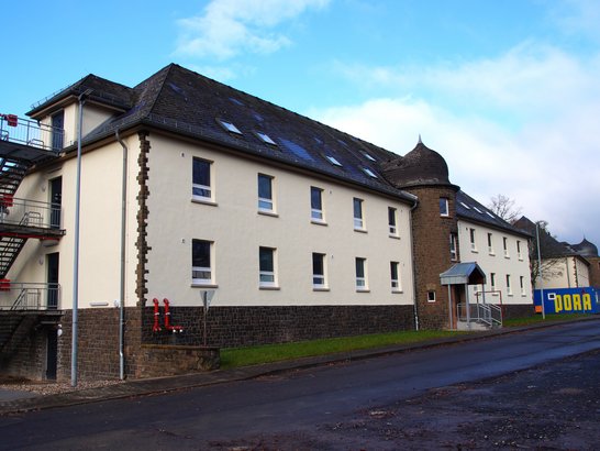 Auf dem Bild ist ein größeres, hell verputztes Gebäude mit dunklem Sockel aus Natur- bzw. Bruchstein zu sehen. Es wirkt funktional und eher sachlich gestaltet. Das Haus hat mehrere Stockwerke und ein geneigtes, dunkel gedecktes Dach mit einigen Dachfenstern. Links am Gebäude befindet sich eine außenliegende Metalltreppe mit roten Geländern, die offenbar als Flucht- oder Zugangstreppe dient. Auf der rechten Seite ist ein runder, turmartiger Gebäudeteil aus dunklerem Stein angebaut, der dem Bau einen leicht historischen oder kasernenartigen Charakter verleiht. Ein kleiner Eingangsbereich mit Vordach ist ebenfalls zu erkennen.