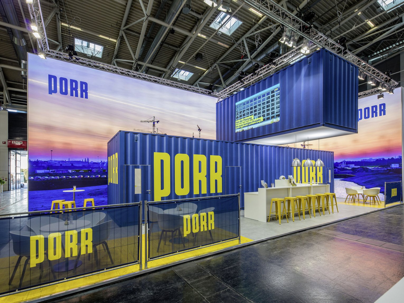 The image shows a modern exhibition stand designed to resemble construction site containers. The containers are painted in a bold blue colour and feature the yellow lettering ‘PORR’ prominently on several sides. Inside the stand is an inviting seating area with tables and chairs, as well as a bar area with several yellow bar stools. A large screen is mounted above the bar, displaying information. The backdrop is an atmospheric cityscape at sunset, which lends the stand additional visual depth and atmosphere.