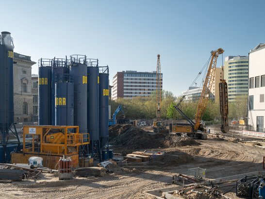 Das Bild zeigt eine Baustelle mit verschiedenen Geräten und Strukturen. Auffällig sind mehrere hohe blaue Silos. Im Vordergrund befindet sich eine große gelbe Baumaschine sowie verstreute Baumaterialien. Im Hintergrund sind Kräne und weitere Baugeräte im Einsatz. Die Baustelle ist von Gebäuden umgeben – links ein historisch wirkendes Gebäude, weiter hinten moderne Bauten. Der Himmel ist klar und blau.