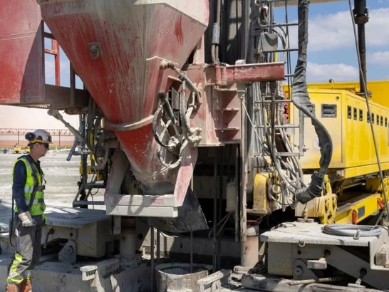 The image shows a construction site. A construction worker stands in front of a large piece of construction equipment.
