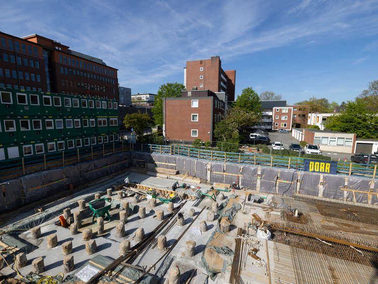 The image shows a large rectangular construction pit surrounded by a construction site fence. Numerous pile heads are exposed at the bottom of the construction pit. Construction equipment and materials can be seen in some places above the piles. In the background are several multi-storey buildings. On the left is a row of green construction containers. A yellow-and-blue flag with the inscription ‘PORR’ is attached to the fence.