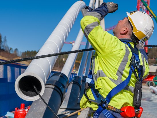 Auf dem Bild ist eine Person bei Arbeiten auf einer großen Baustelle zu sehen. Sie trägt auffällige Schutzkleidung in leuchtendem Gelb, einen Schutzhelm sowie eine persönliche Absturzsicherung mit Gurt und Karabinern. Die Person greift mit beiden Händen nach einem dunklen Seil, das zwischen mehreren großen, hellgrauen Rohren gespannt ist. Die Rohre verlaufen parallel. Im Hintergrund sind weitere Baumaschinen, Hebegeräte und Konstruktionselemente zu erkennen. Der Himmel ist klar und blau.