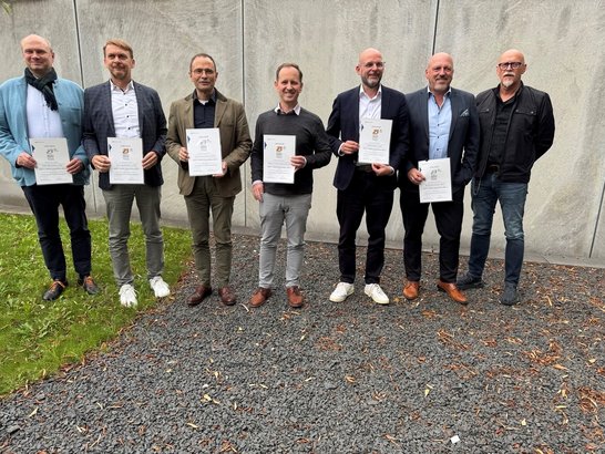 Das Bild zeigt eine Gruppe von sieben Personen, die nebeneinander vor einer grauen Betonwand stehen. Der Untergrund besteht aus dunklem Kies mit einigen verstreuten Blättern, und links ist ein schmaler Streifen grüner Wiese zu sehen. Alle Personen sind formell bis halb-formell gekleidet. Fünf der Personen halten jeweils ein Zertifikat oder eine Urkunde in den Händen. Auf den Dokumenten ist oben ein Wappen oder Logo zu erkennen, darunter Text in schwarzer Schrift.