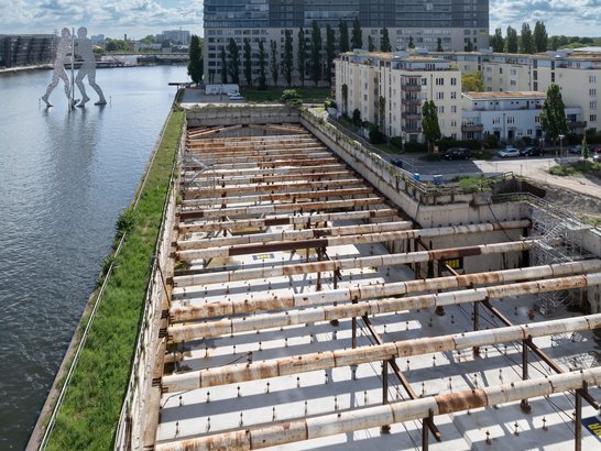 An excavation pit for a construction project on the River Spree in Berlin, featuring a shoring system as well as a view of the Oberbaum Bridge as well as the ‘Molecule Man’ sculpture.
