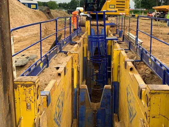 Construction site for pipeline construction with secured trench, excavator, and construction workers in protective clothing; surrounded by trees and construction machinery.