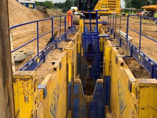 Baustelle im Leitungsbau mit gesichertem Graben, Bagger und Bauarbeitern in Schutzkleidung; umgeben von Bäumen und Baumaschinen.