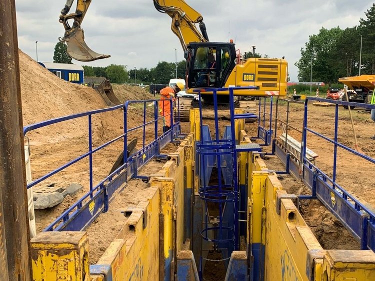 Baustelle im Leitungsbau mit gesichertem Graben, Bagger und Bauarbeitern in Schutzkleidung; umgeben von BĂ€umen und Baumaschinen.