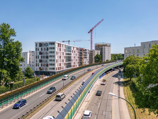 Urban scene with multi-lane road, modern residential buildings and construction cranes on the left, green landscape with trees on the right, as well as cars and vans in sunny weather.
