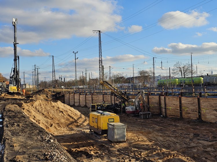 Das Bild zeigt eine große Baugrube im Spezialtiefbau entlang einer Bahnstrecke. Im Vordergrund ist eine tiefe, rechteckige Baugrube mit sandigem Boden zu sehen. Die Baugrubenwände sind mit einer Trägerbohlwand aus horizontal angeordneten Holzbohlen gesichert, die von Stahlträgern gehalten werden. Innerhalb der Baugrube stehen mehrere Baumaschinen, darunter ein gelber Kompressor bzw. ein Aggregat sowie ein Bagger mit Anbaugerät. Links im Bild ragt ein hohes Bohrgerät empor. Entlang der oberen Kante der Baugrube verlaufen Geländer und Absperrungen, die die Baustelle sichern.