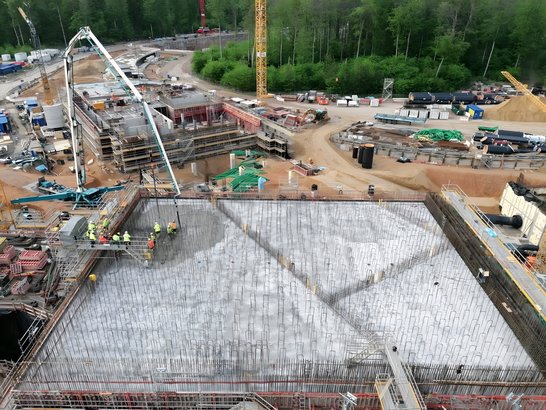 Das Foto zeigt eine großflächige Baustelle inmitten eines bewaldeten Gebiets. Im Vordergrund befindet sich eine massive Betonplatte, die gerade gegossen wird. Aus der Oberfläche ragen zahlreiche Bewehrungsstäbe. Mehrere Bauarbeiter in Sicherheitskleidung sind auf der Platte tätig, während ein Betonpumpenfahrzeug mit langem Ausleger den Beton verteilt. Rund um die Fläche sind komplexe Schalungen und Gerüstkonstruktionen zu erkennen. Im Hintergrund sieht man Kräne, Baumaterialien wie Rohre und Stahlträger sowie eine Zufahrtsstraße. Dahinter erstreckt sich dichter Wald, der die Baustelle umgibt.