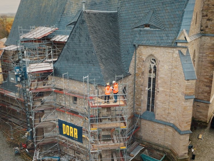 Auf dem Bild ist eine denkmalgeschützte Kirche aus Naturstein zu sehen, die von einem umlaufenden Baugerüst eingerahmt ist. Das Gerüst steht entlang der Fassade und reicht bis unter das steile, schiefergedeckte Kirchendach. An der Außenwand sind typische architektonische Elemente eines spätgotischen Sakralbaus erkennbar, darunter hohe, schmale Fenster und ein massiver Turmbereich. Auf einer Gerüstlage stehen zwei Personen in Warnschutzkleidung, die Arbeiten an der Fassade ausführen. Am Gerüst ist ein Bauschild mit dem Schriftzug „PORR“ angebracht. Der Kirchhof ist teilweise gepflastert; im Hintergrund sind weitere historische Gebäude, parkende Fahrzeuge sowie herbstlich gefärbte Bäume zu sehen.