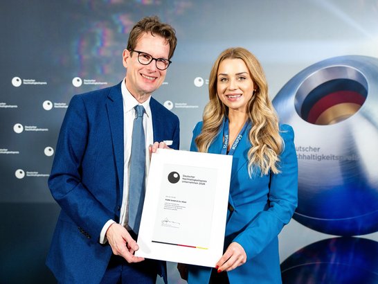 The image shows two people in formal attire, both wearing blue suits. They are holding a large certificate or award together. The certificate features a logo with a black circle and the text ‘German Sustainability Award’, along with other details indicating an award ceremony. In the background is a wall with the German Sustainability Award logo repeated several times and a large, spherical object with coloured segments, which is also part of the brand identity.