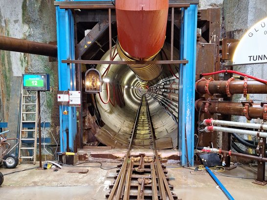 Eingang eines Tunnels mit Schienen im Vordergrund, einem großen gelben und roten Lüftungsschlauch über dem Eingang, mehreren Rohren und Ventilen rechts sowie einer Leiter und einem Bildschirm