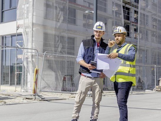 The image shows two construction workers on a building site. They are wearing hard hats and high-visibility vests.