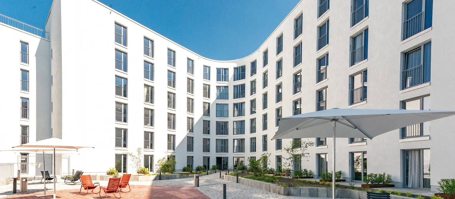 The Fizz -Stresemannquartier, Hamburg The façade with white, floor-to-ceiling windows. In front of this is the inner courtyard with chairs and parasols.