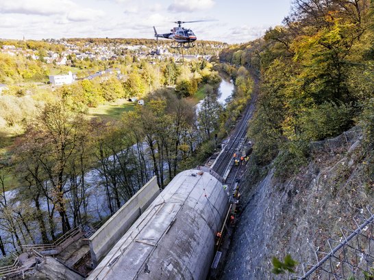 Hubschrauber unterstützt Tunnelbauarbeiten in steilem Gelände und transportiert Material entlang einer Baustelle oberhalb eines Flusses.