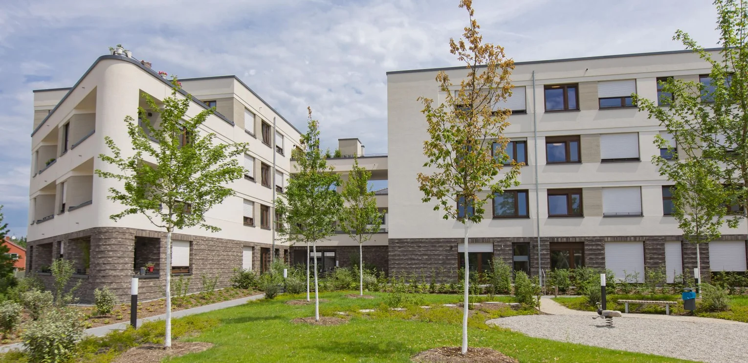 Jackson Three im Franklin Quartier, Mannheim View of the building complex; the ground floor consists of a clinker brick façade; the three remaining upper floors are beige in colour; the building on the left has a triangular shape towards the end of the building; there is a spacious roof terrace on the top floor; the floors below have loggias; several young trees can be seen on the green area in the foreground;