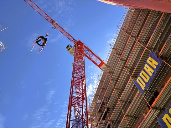 A view from below of a red tower crane with a lifting ring and coloured ribbons, set against a building site covered in scaffolding. PORR banners are displayed on the building’s façade.