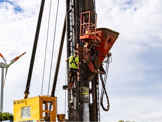 A construction worker in safety clothing operates a large drill with a yellow cabin and orange-red attachment on a construction site. In the background, a wind turbine with orange rotor blade tips stands under a partly cloudy sky.