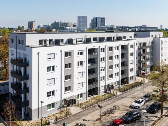 Multi-storey residential building with white-grey façade and balconies in an urban setting, with parked cars, bicycles and green street area – an example of modern urban architecture.