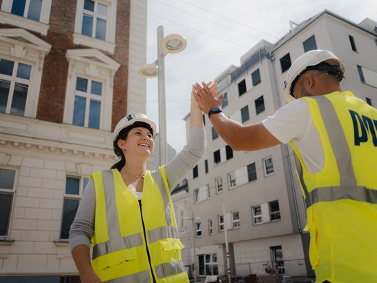 Auf dem Bild sind zwei Personen auf einer Baustelle zu sehen. Beide tragen weiße Schutzhelme und leuchtend gelbe Warnwesten. Sie stehen sich gegenüber und klatschen sich mit den Händen ab. Die Szene spielt im Freien vor mehreren mehrstöckigen Gebäuden: im Hintergrund erkennt man eine Mischung aus älterer Architektur mit Backsteinfassade und neueren, hellen Gebäuden.