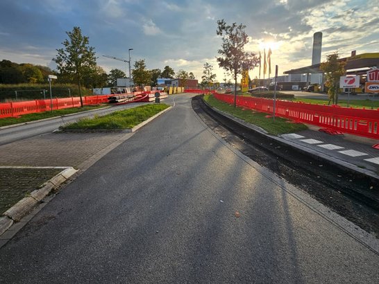 Auf dem Bild sieht man eine leicht geschwungene Straße. Die Fahrbahn ist dunkel asphaltiert und wirkt stellenweise feucht. Beidseitig der Straße stehen auffällige rote Absperrgitter, die auf Baustellenarbeiten hinweisen. Links und rechts der Straße gibt es schmale Grünstreifen. Rechts im Hintergrund sind Industrie- oder Gewerbegebäude zu erkennen, darunter ein Gebäude mit Fahnenmasten und ein hoher, zylindrischer Turm oder Schornstein.