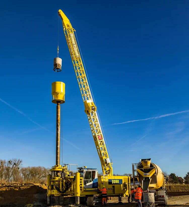 Picture shows a yellow special civil engineering machine. Three construction workers are operating the machine.