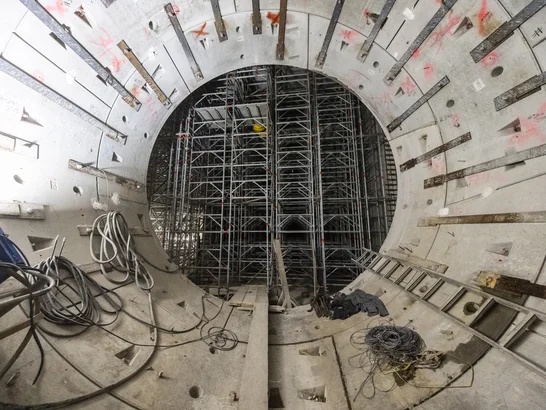 Interior view of a cylindrical structure with concrete walls and extensive scaffolding, cables and tools – showing progress on a tunnel or infrastructure project.