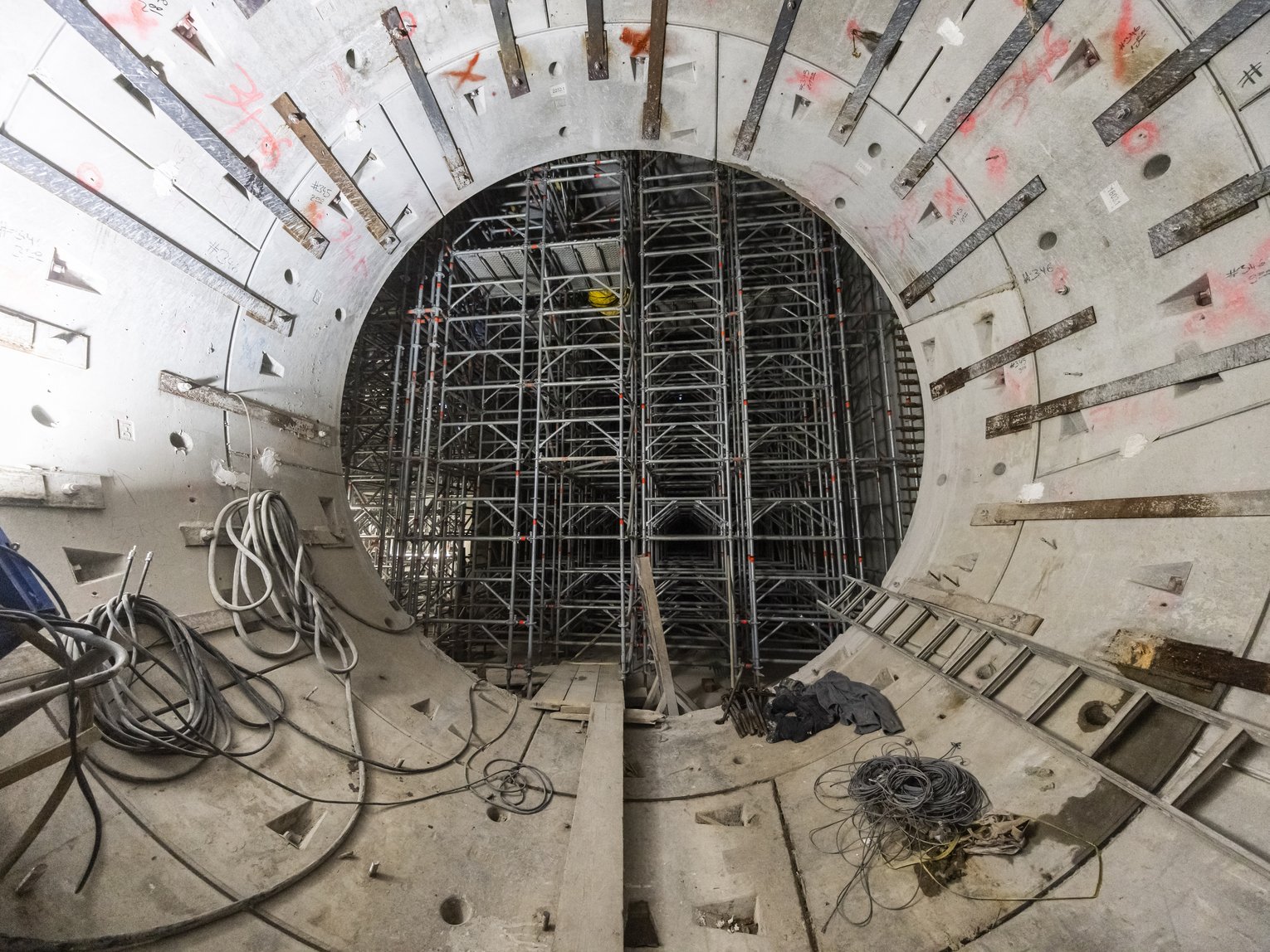  Innenansicht eines zylindrischen Bauwerks mit Betonwänden und umfangreichem Gerüstsystem, Kabeln und Werkzeugen – zeigt den Fortschritt bei einem Tunnel- oder Infrastrukturprojekt.