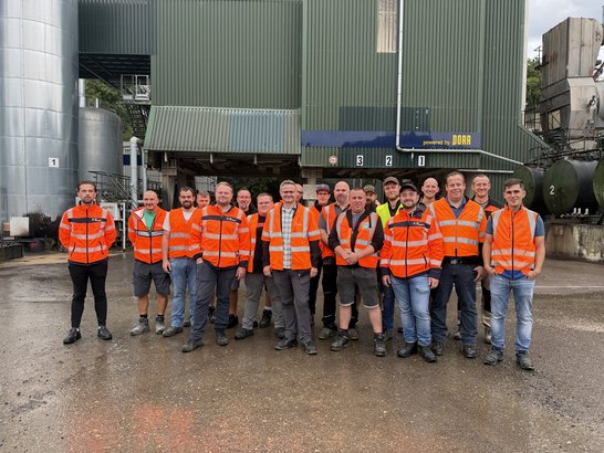 Das Bild zeigt eine Gruppe von Personen, die im Freien vor einer Asphaltmischanlage stehen. Sie tragen orangefarbene Sicherheitswesten sowie unterschiedliche Arbeitskleidung wie Stiefel, Shorts oder lange Hosen. Im Hintergrund sind große Metalltanks, ein grünes Gebäude mit Wellblechverkleidung und Beschilderung sowie weitere industrielle Anlagen zu sehen. Der Boden wirkt nass, was auf kürzlichen Regen hindeutet.