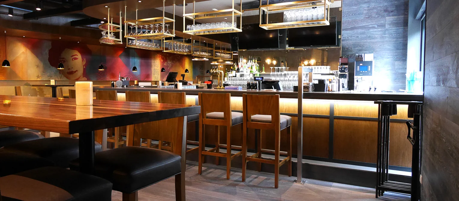 A restaurant for every occasion. Photo: modern wooden furnishings; table with bar stool to the left of the picture; large pictures on the wall in the background; bar with chairs and indirect lighting