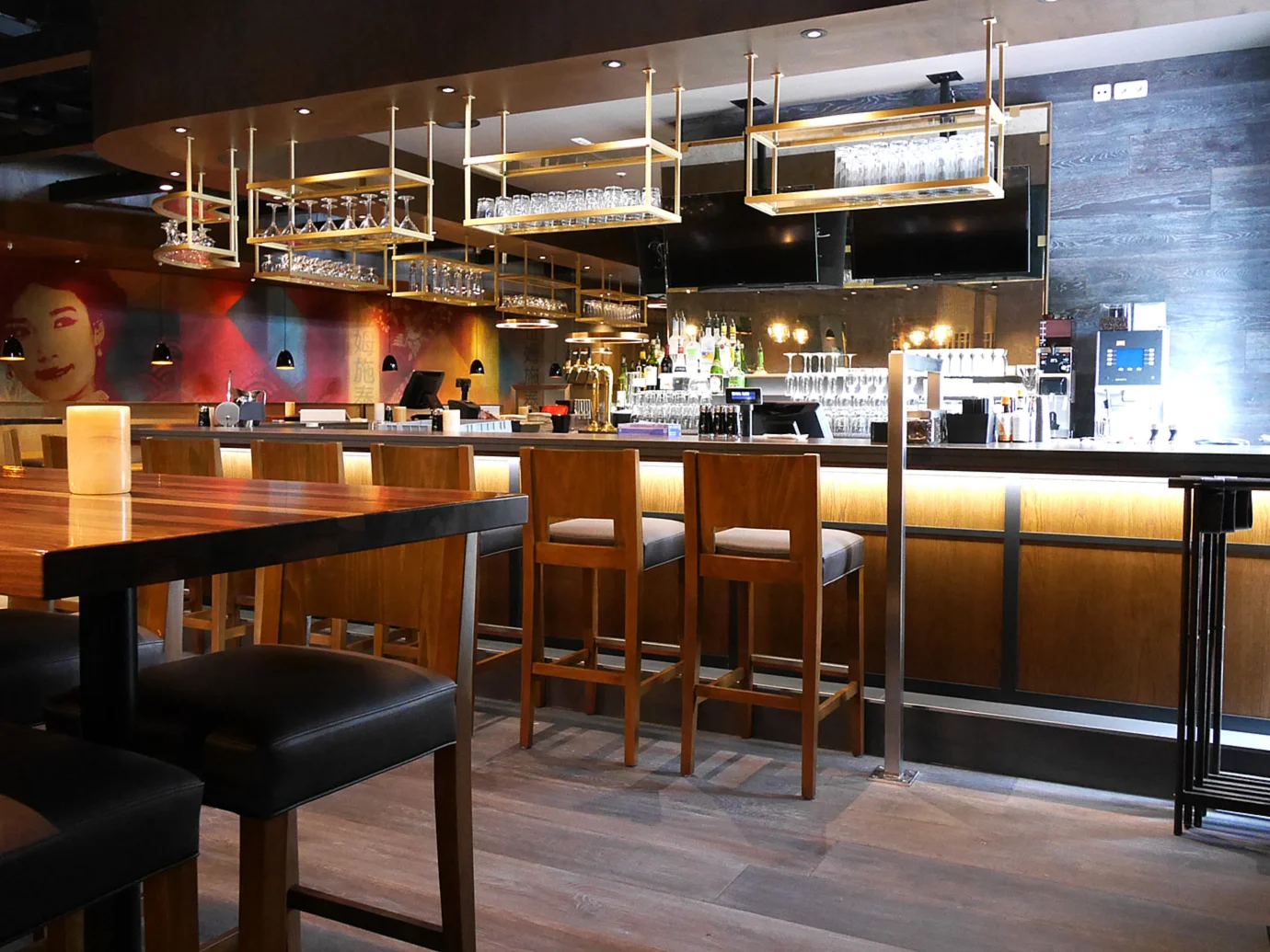A restaurant for every occasion. Photo: modern wooden furnishings; table with bar stool to the left of the picture; large pictures on the wall in the background; bar with chairs and indirect lighting