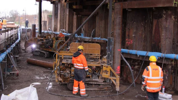 Bild zeigt zwei Bauarbeiter vor einer Ramme unter einen Eisenbahnüberführung; Im Hintergrund arbeiten weitere Personen. Sie arbeiten an einem Stahlrohrpfahl, einem Fertigrammpfahl