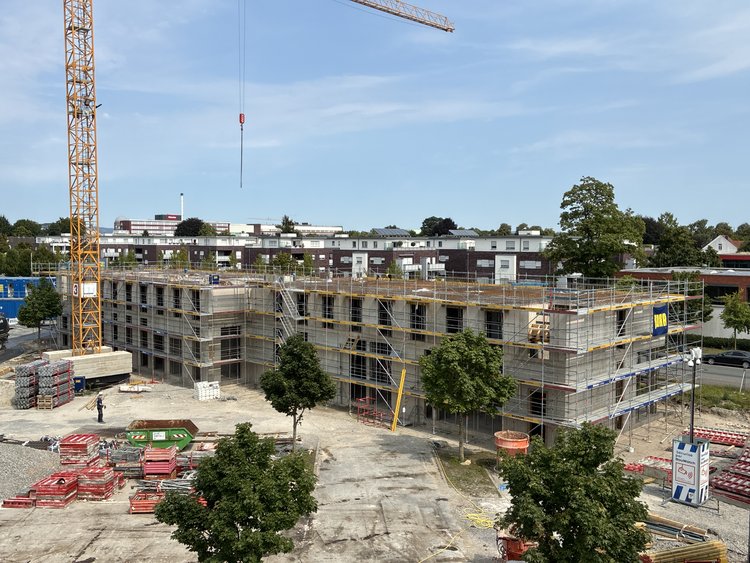 Das Bild zeigt eine Baustelle, auf der ein Gebäude im Bau ist. Das Gebäude ist teilweise fertiggestellt und von Gerüsten umgeben. Auf dem Gelände sind verschiedene Baumaterialien verteilt. Links im Bild befindet sich ein großer gelber Kran. Im Vordergrund sind mehrere Bäume zu sehen, während im Hintergrund Wohngebäude und ein klarer blauer Himmel erkennbar sind.