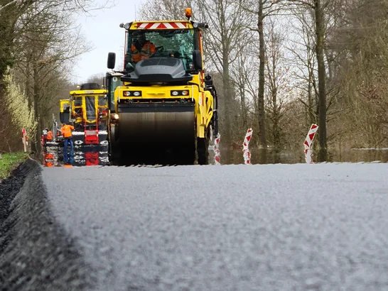 Road construction site on a country road with a yellow roller laying asphalt, surrounded by construction workers, machinery and barriers in a rural setting.