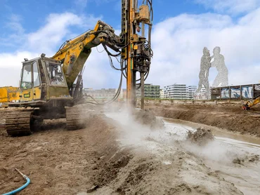 Bild zeigt ein Bohrgerät beim Herstellen einer Düsenstrahlsohle in Berlin; Im Hintergrudn sieht man das bekannte Kunstwerk Berlin und im Vordergrund spritzt der Dreck vom Herstellungsprozess der Dichtsohle. Das Düsenstrahlverfahren in einem Bild