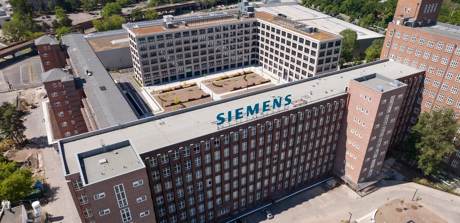 TechnoCampus, Berlin. Building from above; 2 L-shaped buildings form a square; In the foreground is the renovated old building, behind it the new building; On the renovated flat roof is a Siemens logo; In the background you can see the building complex realised by PORR Hochbau.