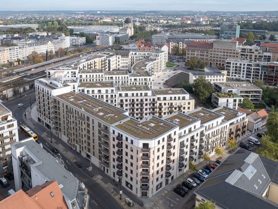 Auf dem Bild ist eine Luftaufnahme eines modernen, blockartigen Wohn- und Geschäftsquartiers in einer städtischen Umgebung zu sehen. Im Vordergrund dominiert ein großer Gebäudekomplex mit mehreren Innenhöfen, der nahezu einen ganzen Straßenblock einnimmt. Die Gebäude sind mehrgeschossig (etwa fünf bis sieben Etagen), mit hellen Fassaden in Beige‑ und Cremetönen. Viele Wohnungen verfügen über Balkone, die regelmäßig angeordnet sind und der Architektur eine klare, rhythmische Struktur geben. Die Flachdächer sind teilweise begrünt.