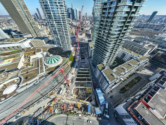 Das Bild zeigt eine dichte, moderne Stadtlandschaft aus der Vogelperspektive: Im Vordergrund ragen mehrere hohe, gläserne Wohn- und Bürohochhäuser in den Himmel. Zwischen zwei besonders markanten Türmen verläuft eine große Baustelle: Man erkennt mehrere rote Baukräne, ein tiefes Baufeld sowie im Bau befindliche Strukturen, die sich längs durch das Areal ziehen. Die Baustelle wirkt wie ein zentrales Entwicklungsprojekt, eingebettet in bestehende Hochhausbebauung. Rund um die Türme sind flachere Gebäude mit begrünten oder technisch genutzten Dächern zu sehen. Teilweise gibt es Dachterrassen, geometrisch angelegte Wege und runde Gebäudeelemente, die einen starken Kontrast zu den kantigen Hochhäusern bilden. Straßen, Gleisanlagen und Verkehrsflächen durchziehen das Quartier und unterstreichen die urbane Dichte.