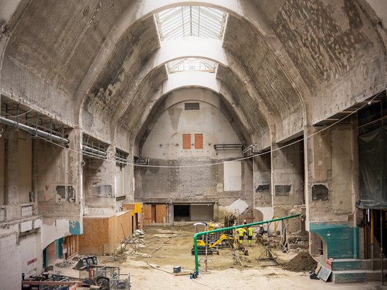 Die Aufnahme zeigt die umfassende Sanierung eines großen historischen Gebäudes mit hohen Gewölbedecken und mehreren Oberlichtern, die den Innenraum mit Tageslicht durchfluten. Im weitläufigen Hallenbereich sind Baugeräte, Materiallager und Baustellenpersonal zu sehen, die an der Modernisierung und Instandsetzung der denkmalgeschützten Bausubstanz arbeiten.