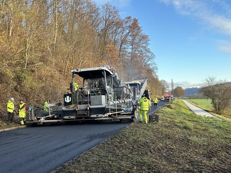The picture shows the asphalt works. You can see a road construction machine and behind it a small forest.
