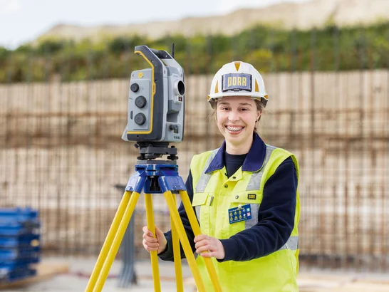 The picture shows a young girl behind a measuring device. She is wearing construction site clothing and a helmet.