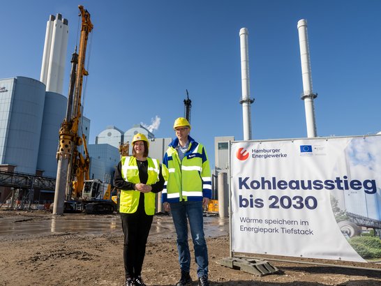 The picture shows a large industrial and construction site scene. In the foreground, two people in protective clothing—wearing high-visibility vests and hard hats—stand on an unpaved, earthen surface. Behind them lies a sprawling industrial site featuring cranes, drilling rigs and several tall, cylindrical chimneys. On the right-hand side of the picture stands a large sign bearing the inscription ‘Phase-out of coal by 2030 – Heat storage at the Tiefstack Energy Park’, accompanied by the logos of Hamburger Energiewerke and the European Union. Modern industrial buildings and technical installations can be seen in the background.