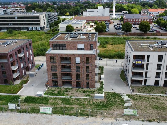 Das Bild zeigt eine Luftaufnahme von drei modernen Wohngebäuden, die in einer Reihe angeordnet sind. Das mittlere Gebäude ist leicht zurückgesetzt im Vergleich zu den beiden anderen. Alle Gebäude haben mehrere Etagen und Flachdächer mit verschiedenen Installationen darauf. Die Fassaden der linken und mittleren Gebäude bestehen aus Backstein, während das rechte Gebäude eine hellere Fassade besitzt.  Rund um die Gebäude befinden sich Wege und Grünflächen. Vor den Gebäuden sind einige Baumaterialien zu sehen, was auf laufende oder kürzlich abgeschlossene Bauarbeiten hindeuten könnte. Im Hintergrund erkennt man weitere Gebäude, Bäume und geparkte Autos.