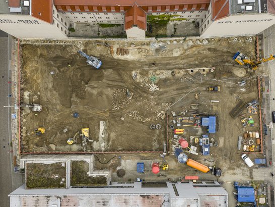 Das Foto zeigt eine große Baustelle aus der Vogelperspektive, die von mehrstöckigen Gebäuden mit roten Dächern umgeben ist. Innerhalb des rechteckigen Geländes wurde der Boden bereits großflächig ausgehoben. Mehrere Baumaschinen sind im Einsatz: Am oberen rechten Rand arbeitet ein großer gelber Bagger, während sich oben links ein weiteres Gerät befindet, vermutlich ein Bohrgerät oder Raupenbagger. Im unteren rechten Bereich sind zahlreiche Materialien und Geräte gelagert, darunter Container, Kabeltrommeln, Rohre sowie ein auffälliger orangefarbener Tank. Im unteren linken Bereich sind kleinere Maschinen und Fahrzeuge zu sehen, die für Erdarbeiten genutzt werden. Der Boden ist uneben und zeigt Spuren von Aushub sowie teilweise sichtbare Fundamentreste.